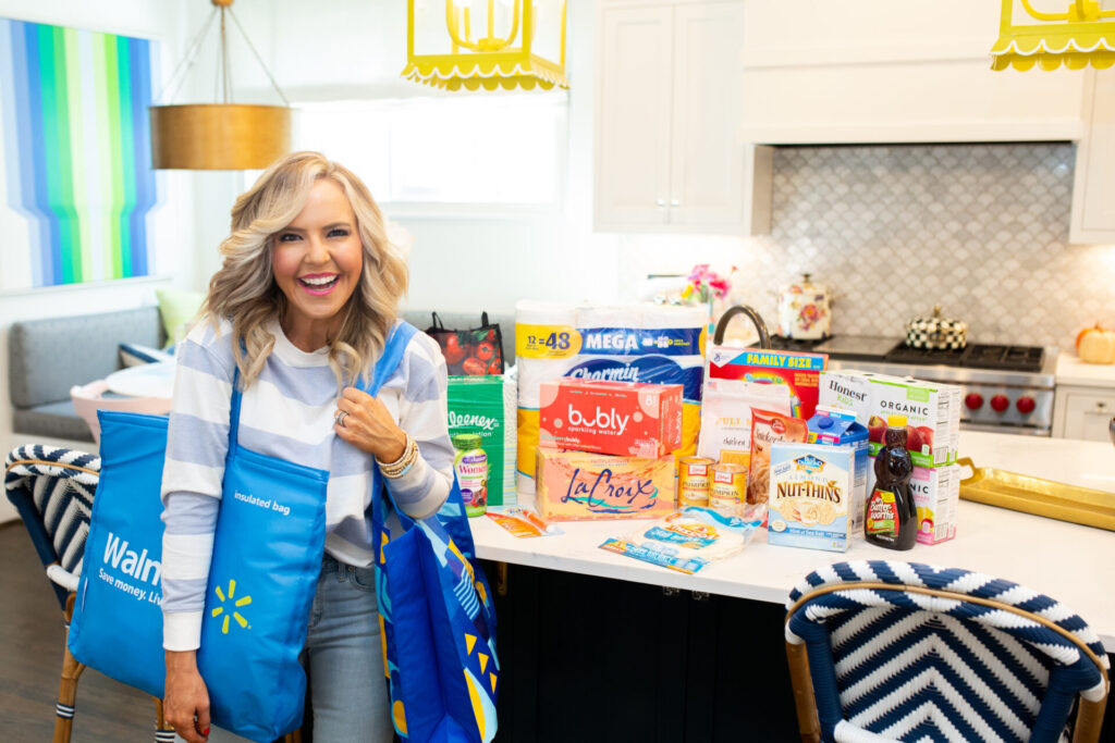 Walmart Membership by popular Nashville lifestyle blog, Hello Happiness: image of Natasha Stoneking holding re-usable Walmart shopping bags and standing next to her kitchen island that's covered with Kleenex, Bubly, La Croix, Mission tortillas, Charmin mega roll toilet paper, Betty Crocker snickerdoodle mix, Almond things, Libby's canned pumpkin, Mrs. ButterWorth's syrup, Lucky Charms cereal and Tide to-go stick. 