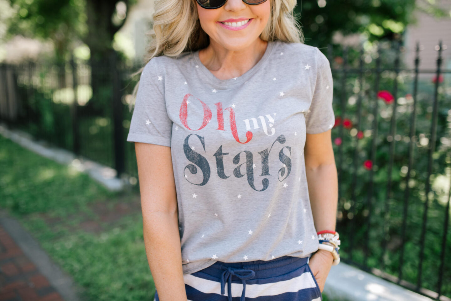 4th of July Outfits by popular Nashville fashion blog, Hello Happiness: image of a woman standing outside on a red brick walkway and wearing a grey Oh My Stars t-shrit with blue and white stripe shorts, sliver and white sneakers and holding a grey and white checkered handbag.