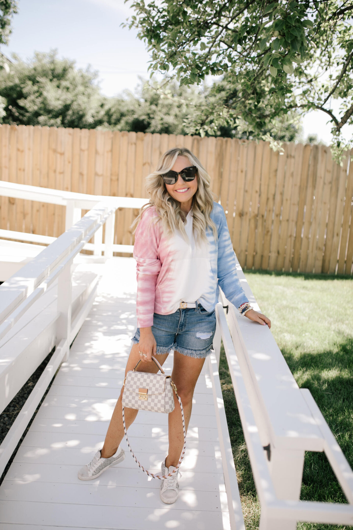 4th of July Outfits by popular Nashville fashion blog, Hello Happiness: image of a woman wearing a red, white, and blue tie dye hoodie, distress hem denim shorts, white Louis Vuitton belt, silver and white sneakers, black frame sunglasses, and holding a grey and white checkered handbag. 