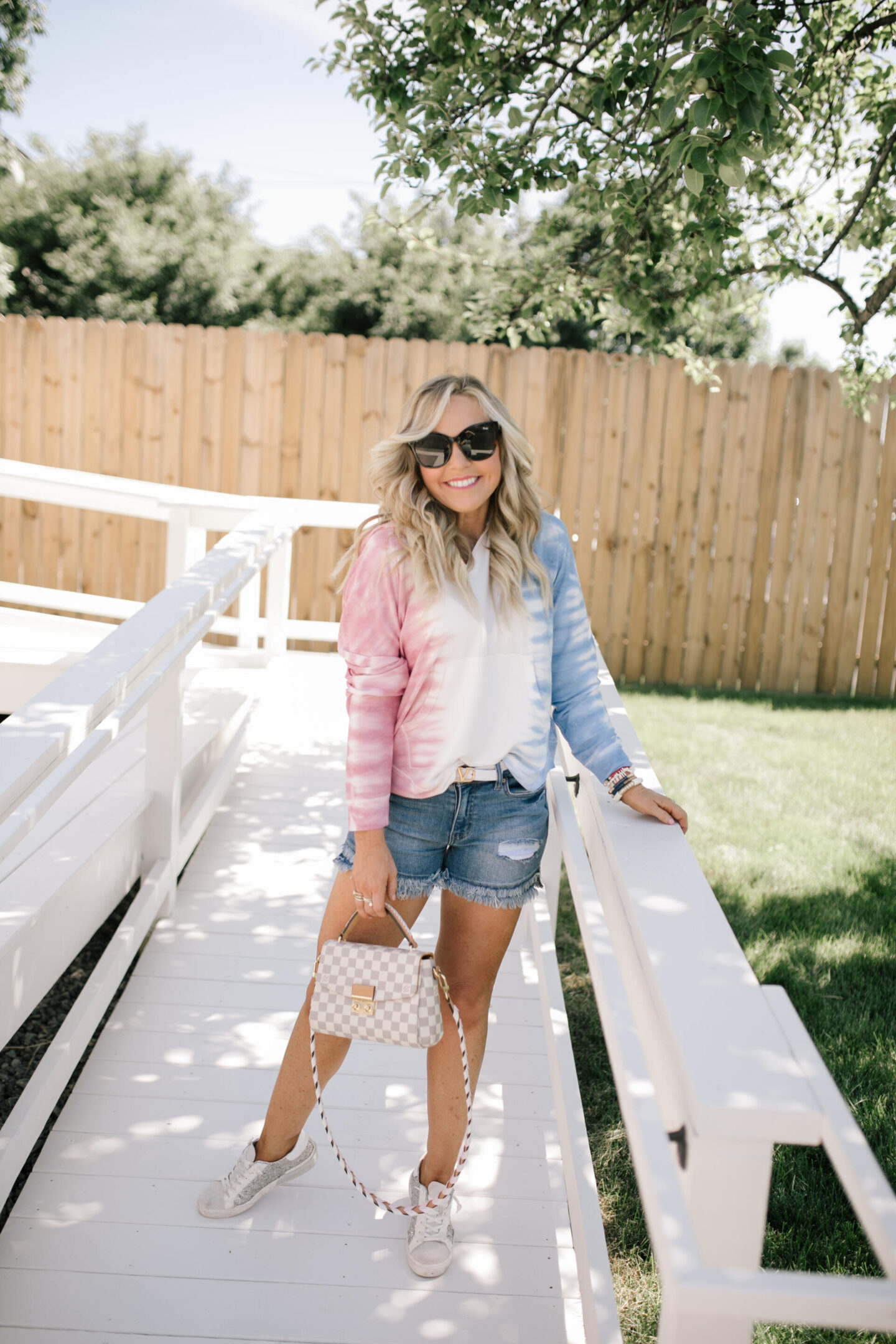 4th of July Outfits by popular Nashville fashion blog, Hello Happiness: image of a woman wearing a red, white, and blue tie dye hoodie, distress hem denim shorts, white Louis Vuitton belt, silver and white sneakers, black frame sunglasses, and holding a grey and white checkered handbag.