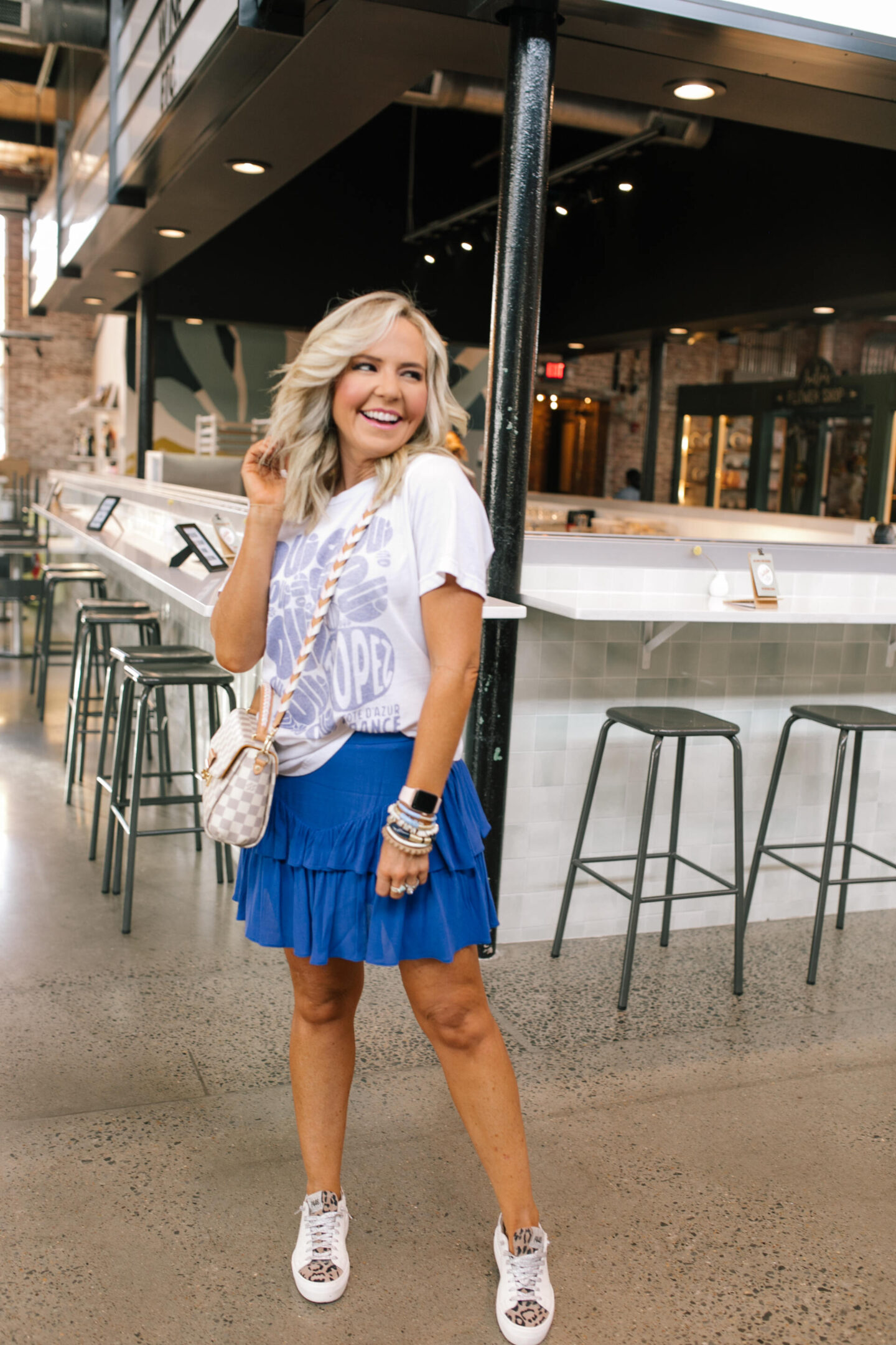 Revolve Fashion by popular Nashville fashion blog, Hello Happiness: image of a woman wearing a blue and white graphic t-shirt with a royal blue tiered mini skirt, stack bracelets, and leopard print sneakers.