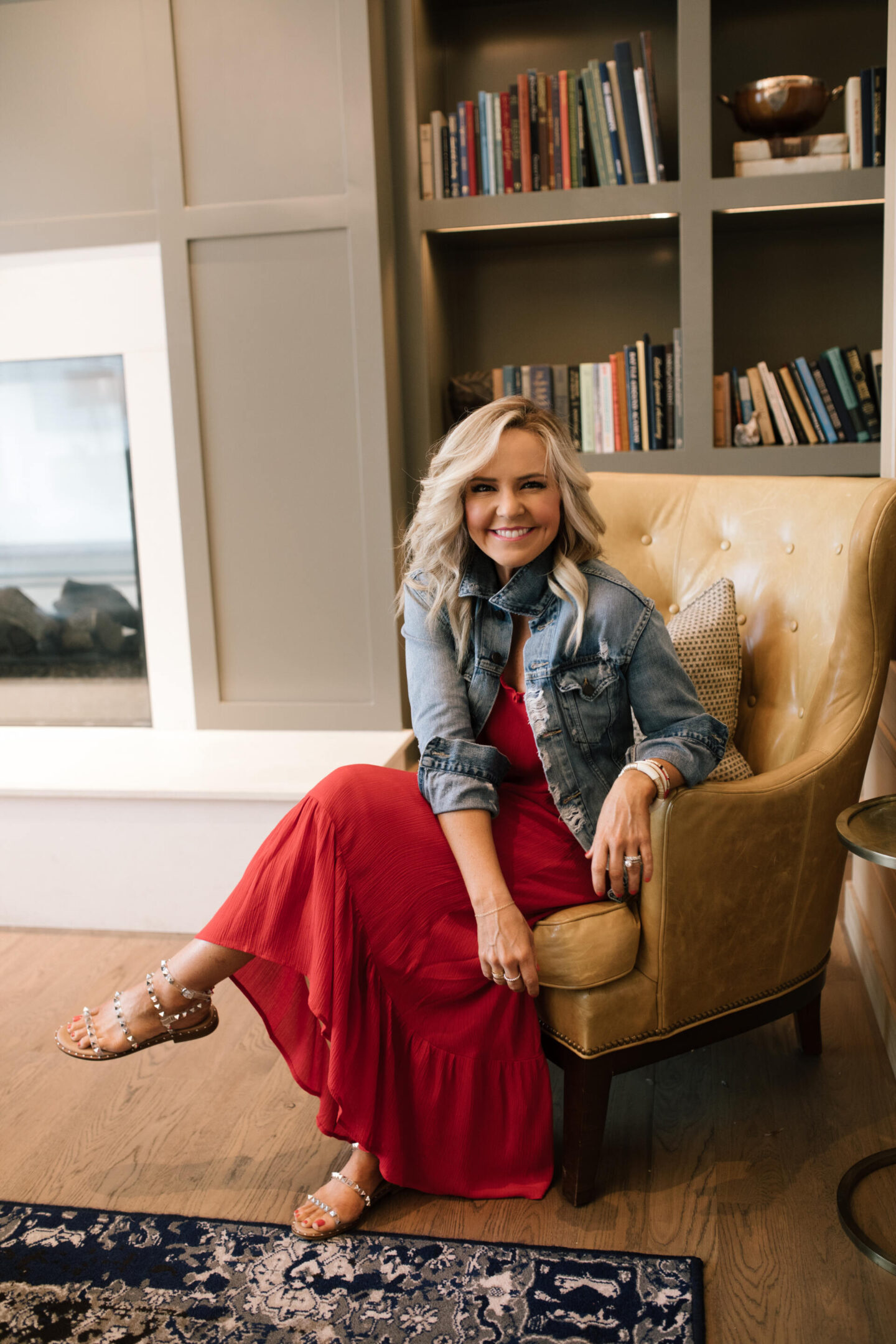 Harpeth Hotel by popular Nashville travel blog, Hello Happiness: image of Natasha Stoneking wearing a red maxi dress, denim jacket and clear studded strap sandals while sitting in a light brown leather chair. 