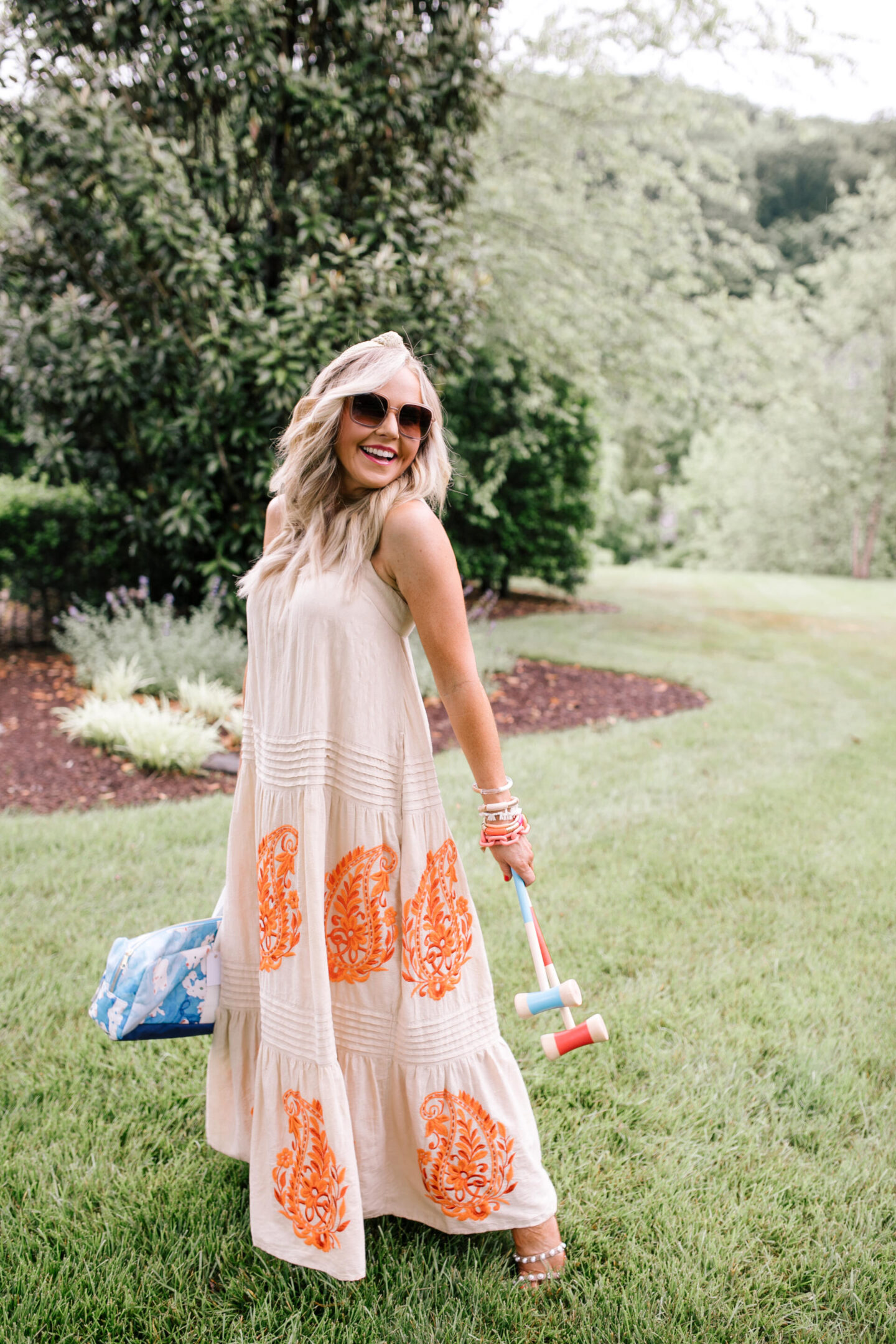 Anthropologie Products by popular Nashville life and style blog, Hello Happiness: image of Natasha Stoneking standing outside and holding some crochet mallets while wearing a pink maxi dress with orange embroidery and a pair of sunglasses. 