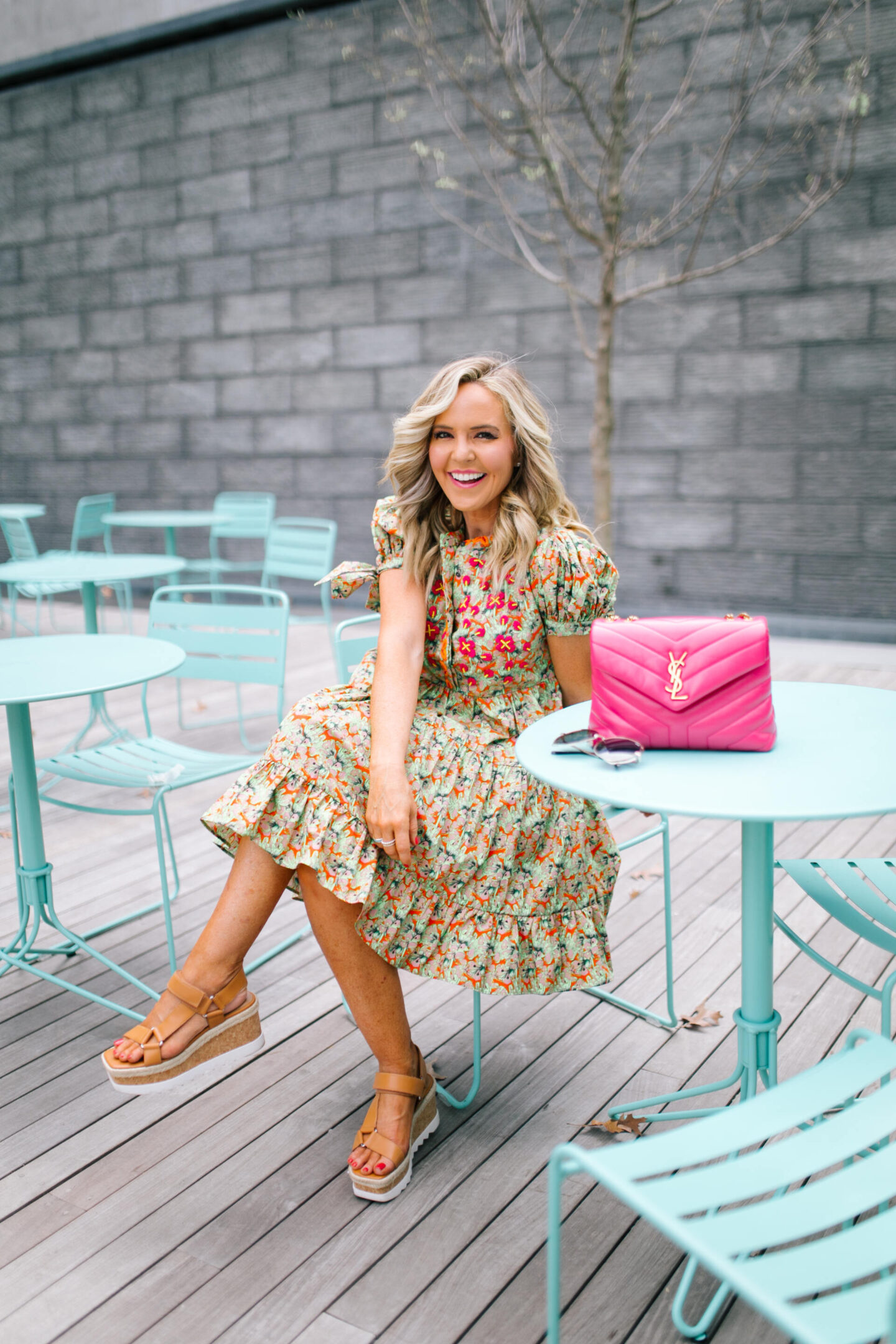 Anthropologie Sale by popular Nashville life and style blog, Hello Happiness: image of Natasha Stoneking sitting at a blue outdoor table set and wearing a green floral print dress, camel color strap platform wedge sandals, and a pink YSL bag. 