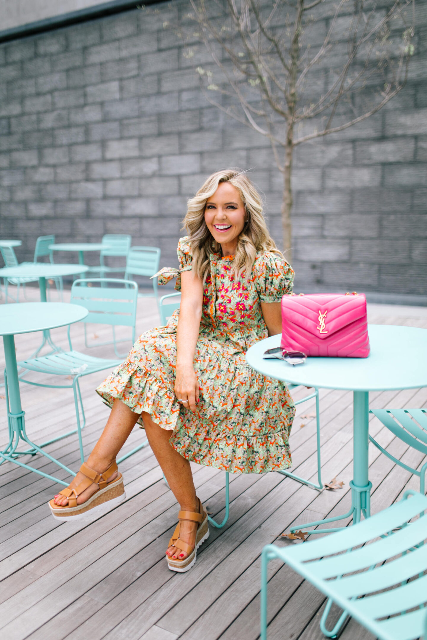 Anthropologie Sale by popular Nashville life and style blog, Hello Happiness: image of Natasha Stoneking sitting at a blue outdoor table set and wearing a green floral print dress, camel color strap platform wedge sandals, and a pink YSL bag.