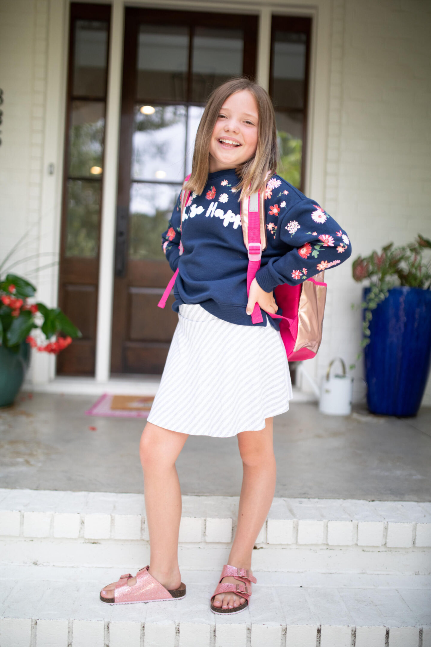 Back to School Essentials by popular Nashville lifestyle blog, Hello Happiness: image of a young girl wearing a blue floral print sweatshirt, grey and white stripe skirt, gold and pink backpack, and shimmer strap sandals. 