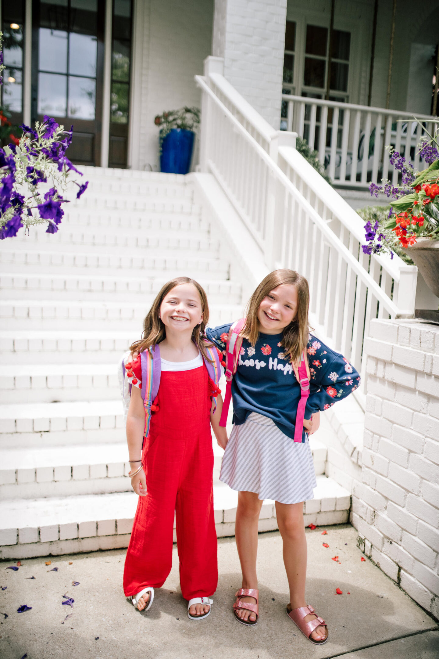 Back to School Essentials by popular Nashville lifestyle blog, Hello Happiness: image of two young girls standing together and wearing a blue floral print sweatshirt, grey and white stripe skirt, gold and pink backpack, shimmer strap sandals, red and white pom pom jumpsuit, rainbow backpack and white strap sandals.