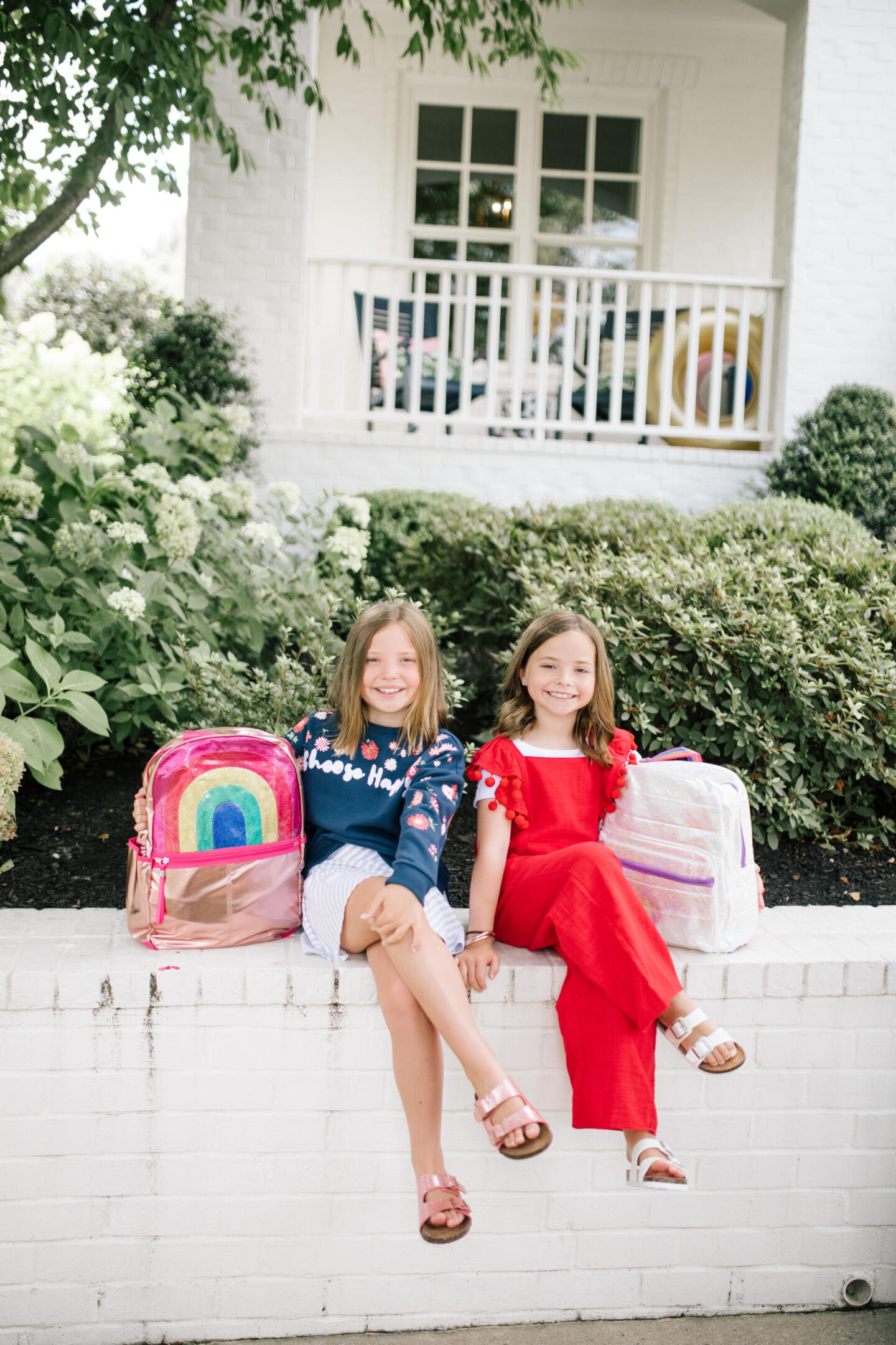 Back to School Essentials by popular Nashville lifestyle blog, Hello Happiness: image of two young girls standing together and wearing a blue floral print sweatshirt, grey and white stripe skirt, gold and pink backpack, shimmer strap sandals, red and white pom pom jumpsuit, rainbow backpack and white strap sandals.