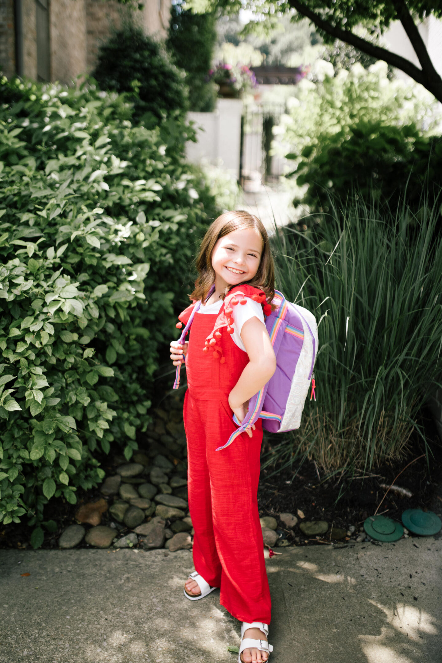 Back to School Essentials by popular Nashville lifestyle blog, Hello Happiness: image of a young girl wearing a red and white pom pom jumpsuit, rainbow backpack and white strap sandals. 