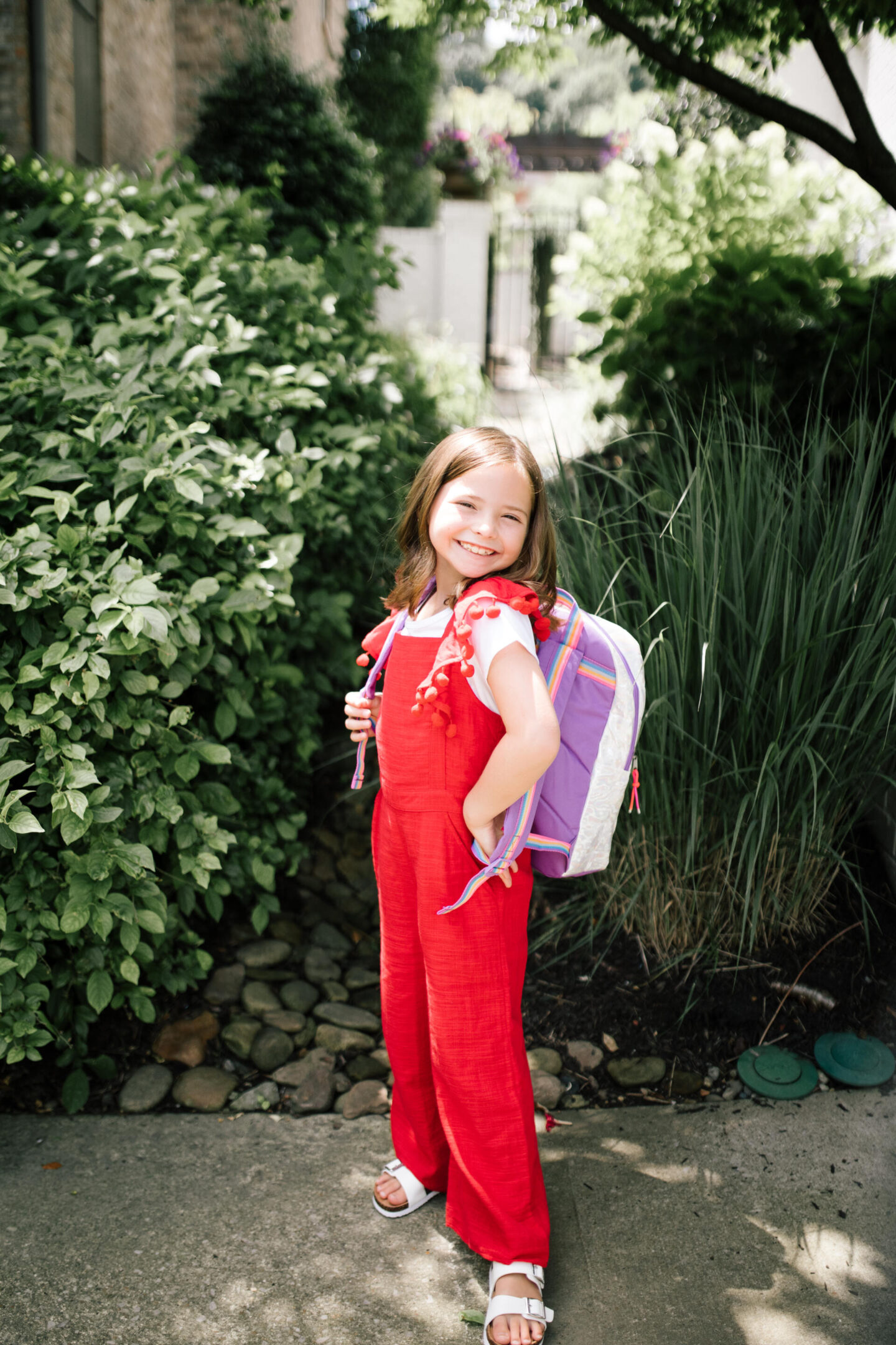 Back to School Essentials by popular Nashville lifestyle blog, Hello Happiness: image of a young girl wearing a red and white pom pom jumpsuit, rainbow backpack and white strap sandals.