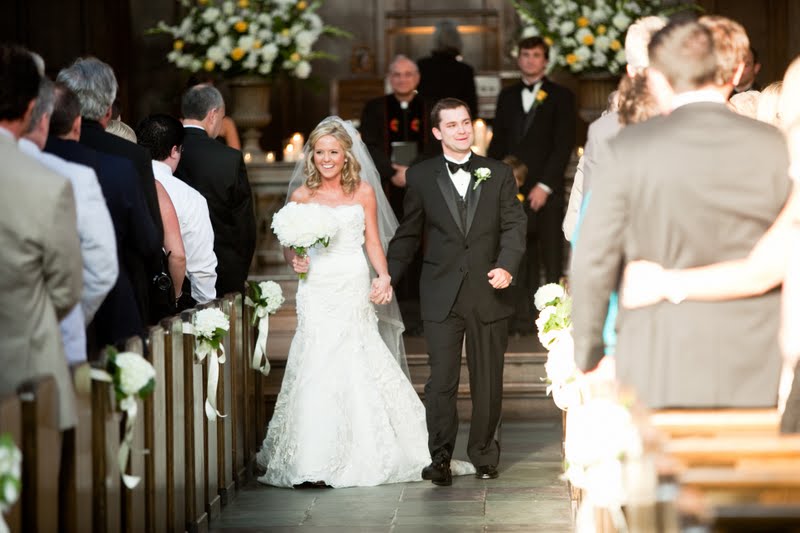 10 Year Wedding Anniversary by popular Nashville lifestyle blog, Hello Happiness: image of a bride wearing a strapless lace wedding dress and holding a white floral bouquet and a groom wearing a black tuxedo and holding hands as they walk down the aisle inside a cathedral.